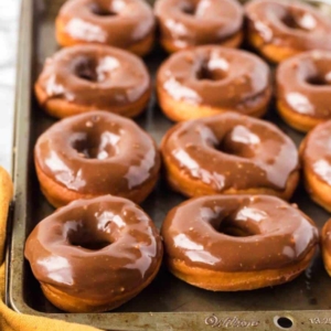 A baking tray filled with freshly glazed chocolate donuts, arranged in rows. A yellow cloth is partially visible in the bottom left corner.