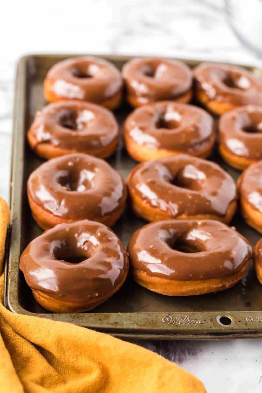 A baking tray filled with freshly glazed chocolate donuts, arranged in rows. A yellow cloth is partially visible in the bottom left corner.