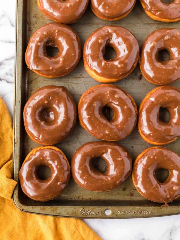 A baking tray filled with twelve glazed chocolate donuts sits on a white marble surface, with a yellow cloth partially visible in the bottom left corner.