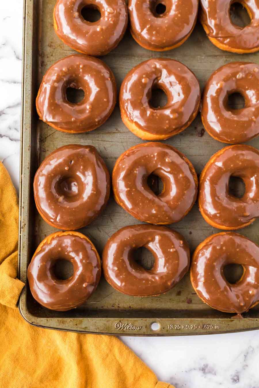 A baking tray filled with twelve glazed chocolate donuts sits on a white marble surface, with a yellow cloth partially visible in the bottom left corner.