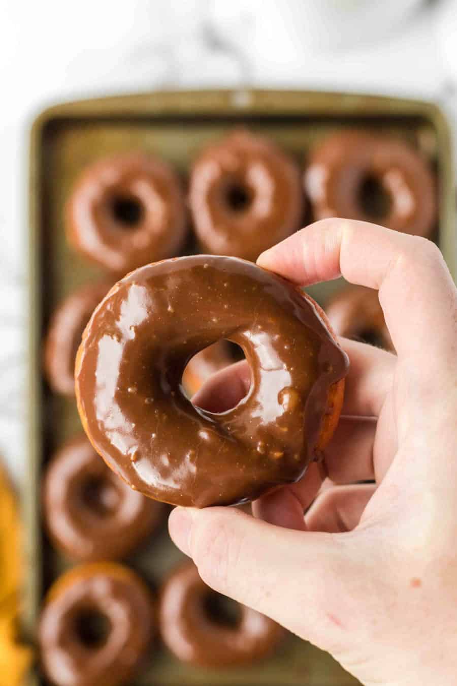 A hand holds up a chocolate-glazed donut in front of a tray filled with more chocolate-glazed donuts in the background.