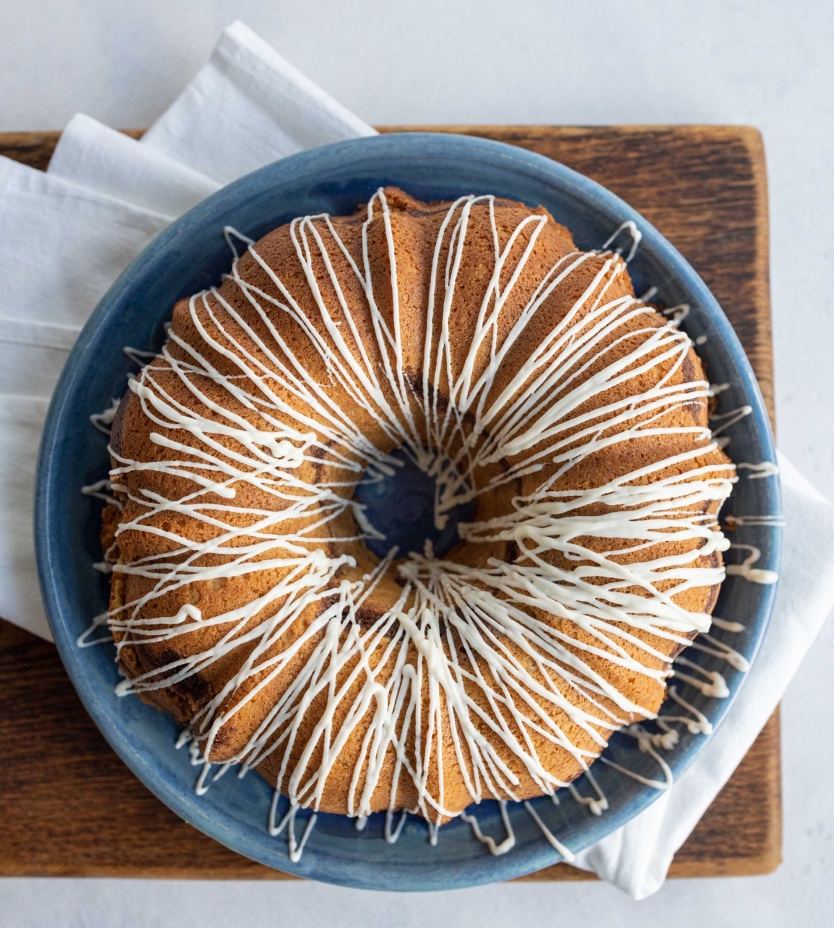 A round bundt cake with white icing drizzled in a zigzag pattern sits on a blue plate, placed on a wooden board with a white napkin underneath.
