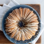 A round bundt cake with white icing drizzled in a zigzag pattern sits on a blue plate, placed on a wooden board with a white napkin underneath.