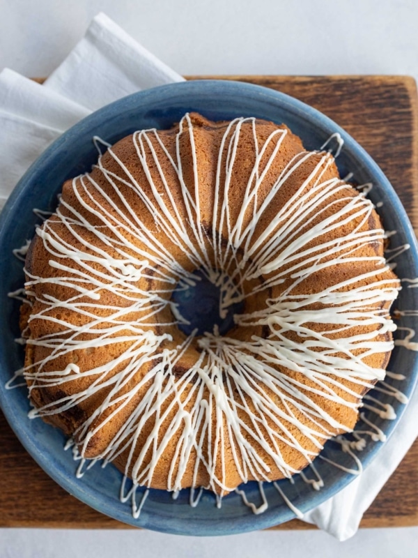 A round bundt cake with white icing drizzled in a zigzag pattern sits on a blue plate, placed on a wooden board with a white napkin underneath.
