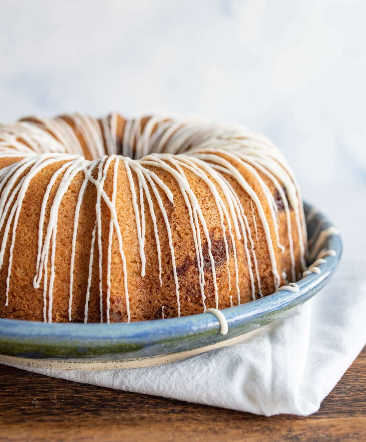 A bundt cake with a golden-brown crust is drizzled with white icing, sitting on a blue ceramic plate atop a folded white cloth on a wooden surface.