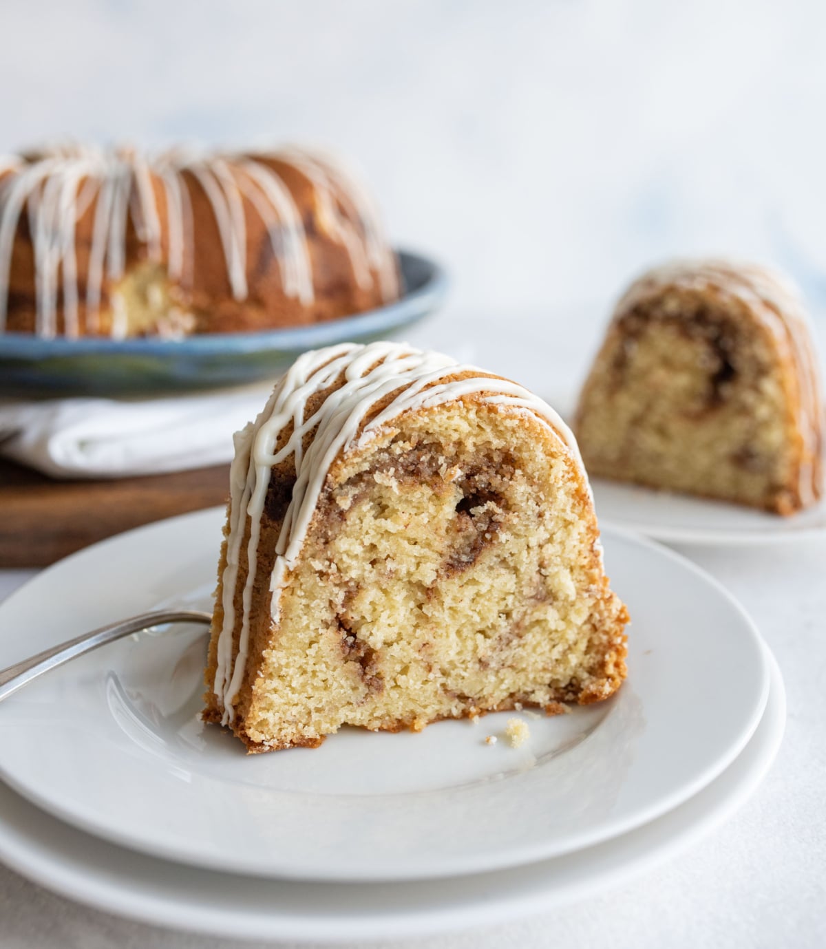 A slice of bundt cake with cinnamon swirl and white icing drizzle sits on a white plate with a fork. More cake is visible in the background on a serving platter and another plate.
