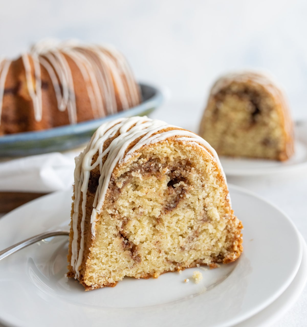 A close-up of a slice of cinnamon swirl bundt cake with white icing drizzle, served on a white plate with a fork. More cake slices are visible in the background.
