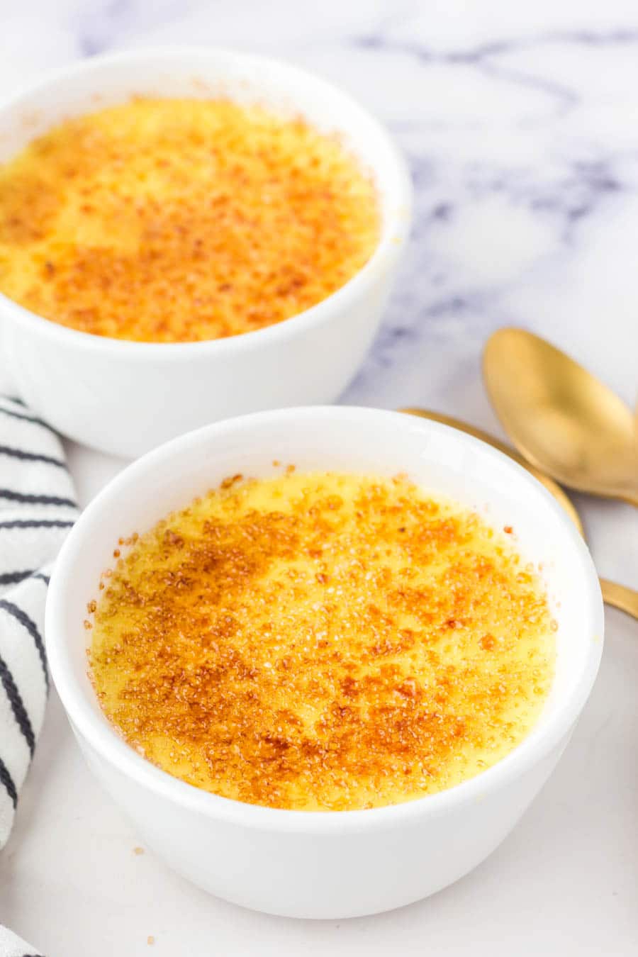 Two white ramekins filled with cr&egrave;me br&ucirc;l&eacute;e topped with a golden, caramelized sugar crust, placed on a white surface with two gold spoons and a striped cloth nearby.