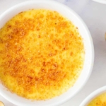 A close-up of crème brûlée in a white ramekin, showing its caramelized, golden-brown sugar crust on top. Other ramekins and a striped napkin are partially visible in the background.