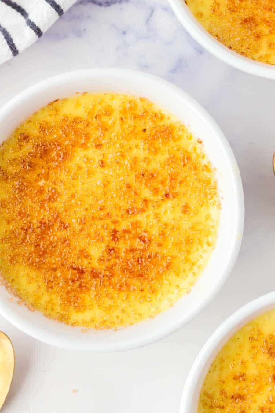 A close-up of crème brûlée in a white ramekin, showing its caramelized, golden-brown sugar crust on top. Other ramekins and a striped napkin are partially visible in the background.