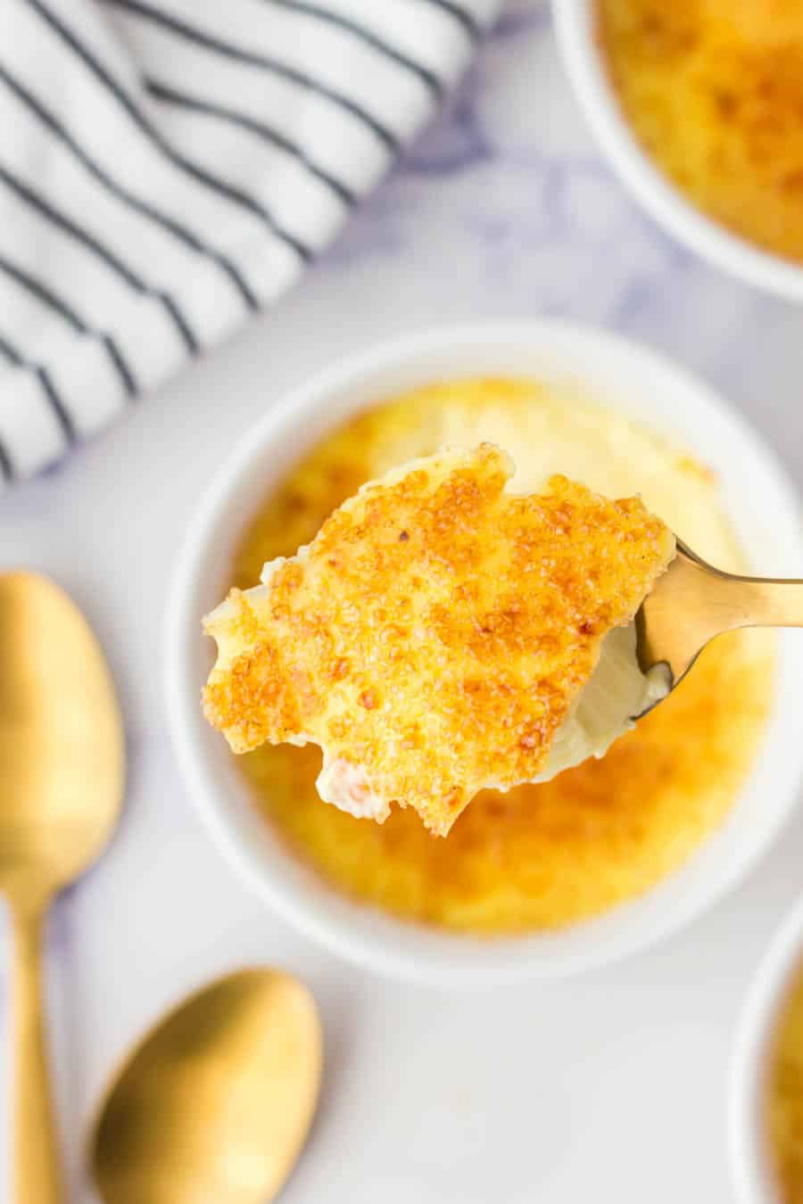 A spoonful of cr&egrave;me br&ucirc;l&eacute;e with a caramelized sugar crust is held above a ramekin, with gold spoons and a striped napkin nearby on a white surface.