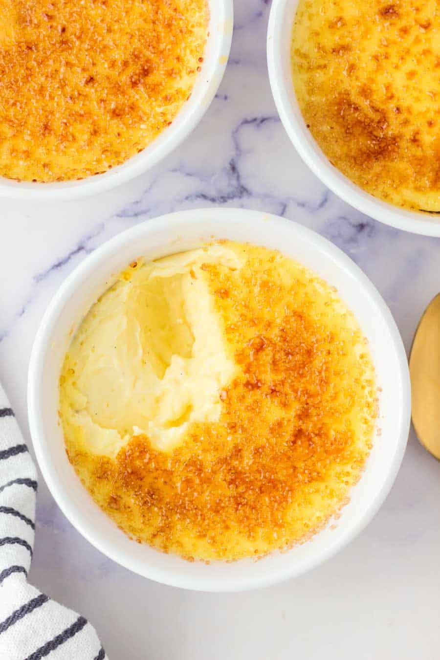 A round white ramekin filled with creamy cr&egrave;me br&ucirc;l&eacute;e, topped with a golden, caramelized sugar crust, and a spoonful removed to reveal the smooth custard inside. Other ramekins and a striped cloth are nearby.