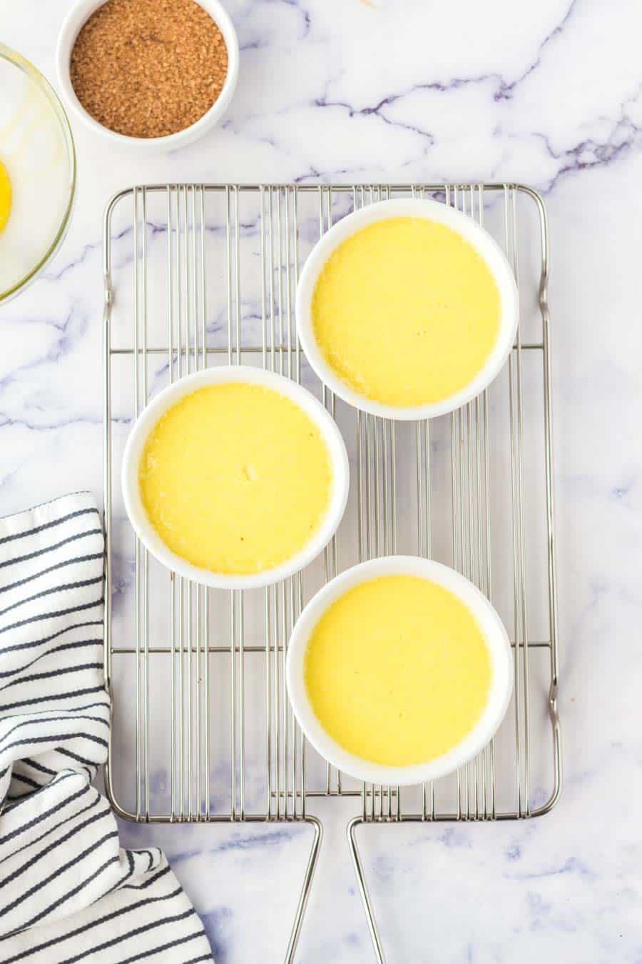 Three small white ramekins filled with a yellow custard sit on a wire cooling rack. A bowl of brown sugar and a striped kitchen towel are nearby, on a white marble surface.