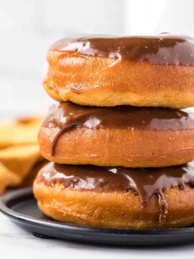 Two chocolate-glazed donuts stacked on a black plate, with another donut partially visible in the background. The donuts have a shiny chocolate topping and golden-brown, fluffy texture.