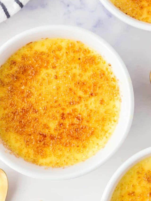 A close-up of a white ramekin filled with cr&egrave;me br&ucirc;l&eacute;e, featuring a caramelized golden-brown sugar crust on top, set on a light surface with a partial view of other ramekins nearby.