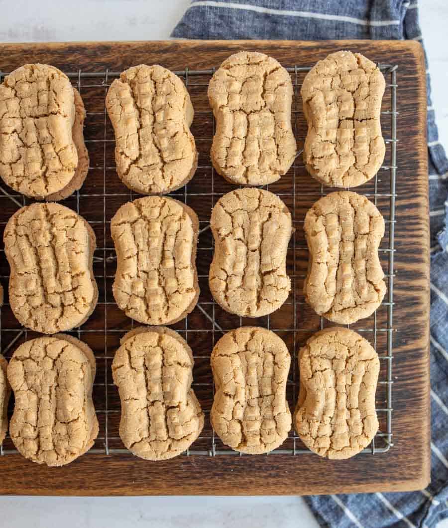 A batch of oval, peanut-shaped cookies with a crisscross pattern rest on a cooling rack placed on a wooden board, with a striped cloth partially visible underneath.