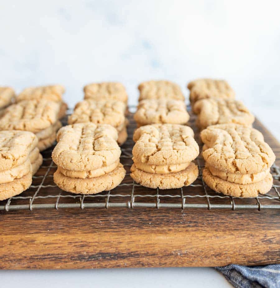 Rows of peanut butter sandwich cookies with creamy filling are arranged on a wire cooling rack set atop a wooden board. The cookies have a crisscross pattern on top.