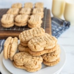 A plate of homemade peanut butter sandwich cookies sits in the foreground, with more cookies cooling on a wire rack and two glasses of milk in the background. A blue and white cloth is underneath.
