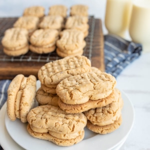 A plate of homemade peanut butter sandwich cookies sits in the foreground, with more cookies cooling on a wire rack and two glasses of milk in the background. A blue and white cloth is underneath.
