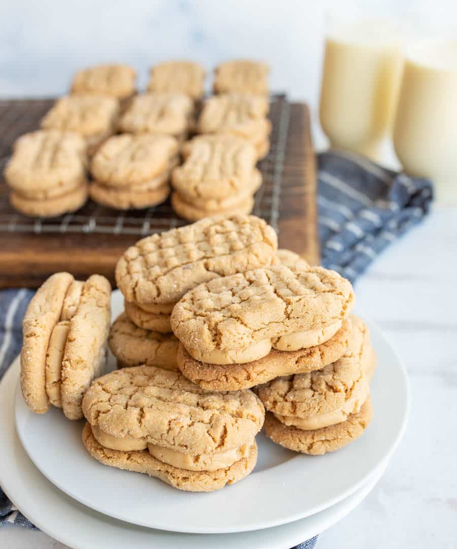 A plate of homemade peanut butter sandwich cookies sits in the foreground, with more cookies cooling on a wire rack and two glasses of milk in the background. A blue and white cloth is underneath.