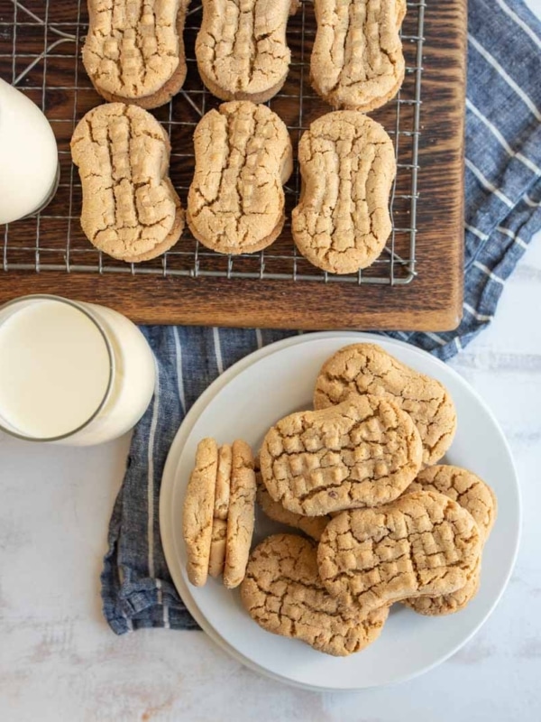 A plate and a cooling rack with peanut butter cookies, alongside two glasses of milk on a blue striped cloth.