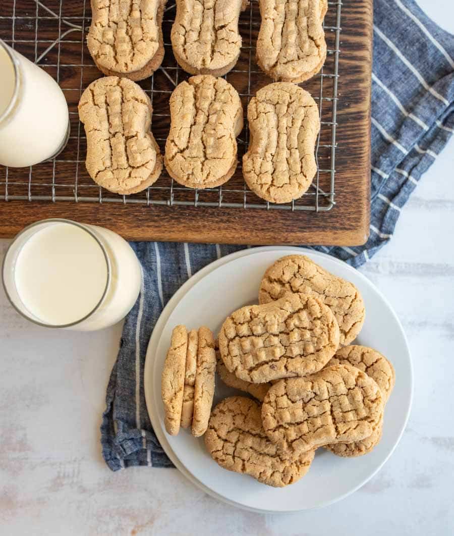 A plate and a cooling rack with peanut butter cookies, alongside two glasses of milk on a blue striped cloth.