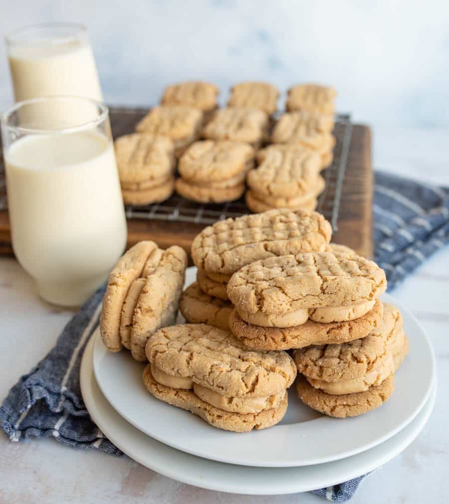 A plate of peanut butter sandwich cookies sits in front of two glasses of milk, with more cookies cooling on a wire rack in the background. The cookies have a crisscross pattern and creamy filling.
