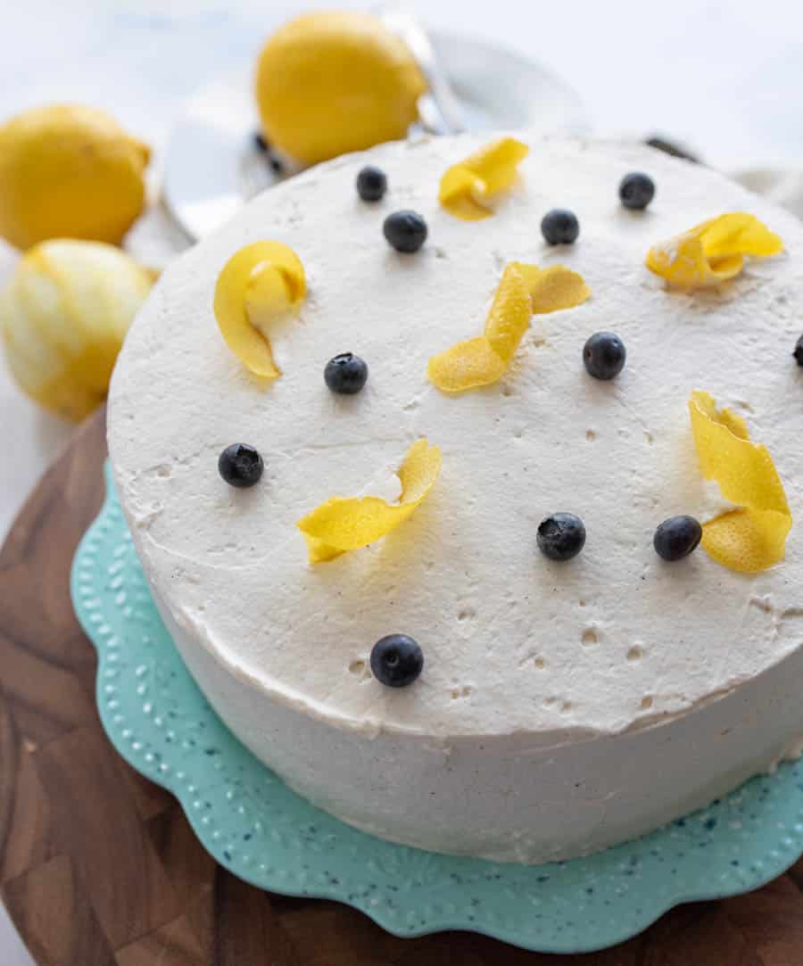 A round cake with white frosting, decorated with fresh blueberries and lemon peel curls, sits on a turquoise plate. Lemons are in the background on a wooden surface.