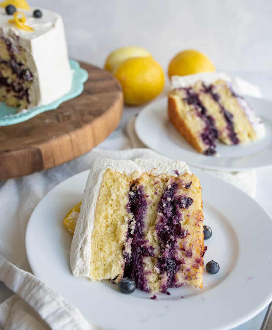 A slice of lemon blueberry cake with white frosting sits on a white plate, garnished with blueberries and a lemon wedge. More cake slices and whole lemons are visible in the background.