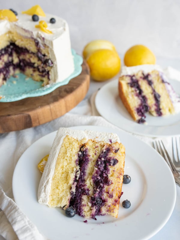 A slice of lemon blueberry layer cake with white frosting is served on a plate, with whole blueberries scattered around. The rest of the cake, partially sliced, sits on a cake stand in the background. Lemons are also visible nearby.
