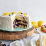 A round lemon and blueberry layered cake with white frosting sits on a wooden cake stand. A large slice is missing, revealing the purple blueberry filling. Lemon peels and a few blueberries decorate the top. Lemons are in the background.
