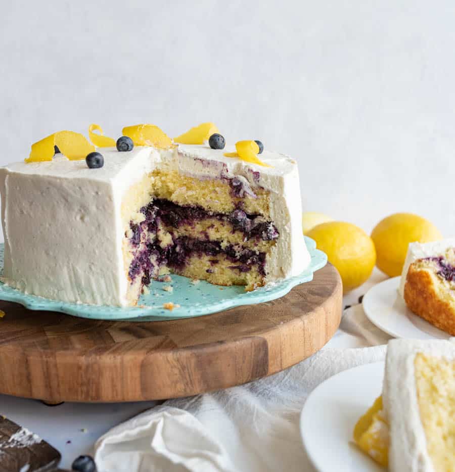 A round lemon and blueberry layered cake with white frosting sits on a wooden cake stand. A large slice is missing, revealing the purple blueberry filling. Lemon peels and a few blueberries decorate the top. Lemons are in the background.