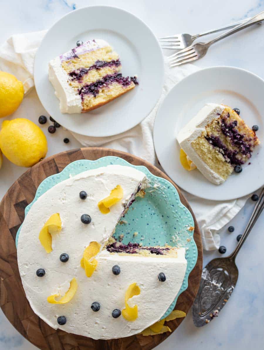 A lemon blueberry layer cake with white frosting, lemon peel curls, and blueberries on top. One slice is missing and served on a plate, with two more plated slices nearby. Lemons and blueberries are scattered around.