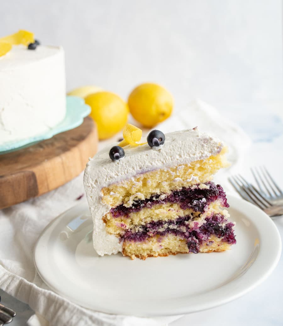 A slice of layered lemon cake with white frosting and blueberry filling is on a white plate. The slice is garnished with blueberries and a lemon twist. The whole cake and fresh lemons are in the background.