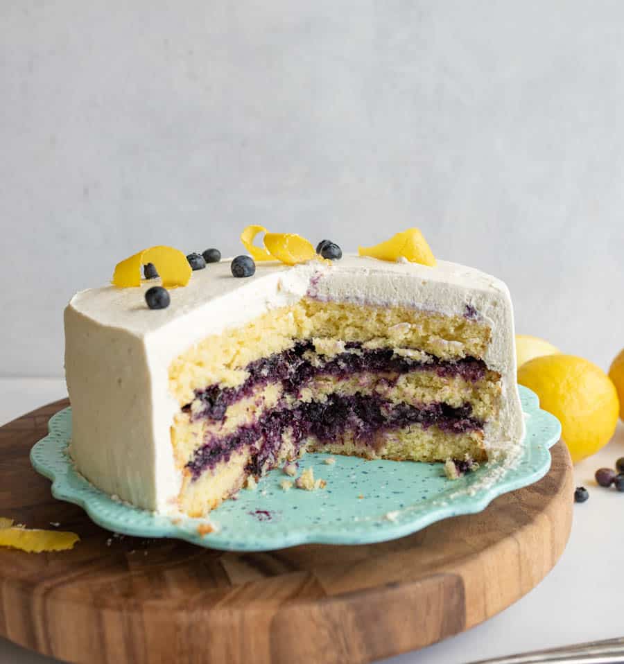 A partially sliced layered cake with white frosting and blueberry filling sits on a blue plate. The cake is decorated with lemon peels and blueberries, with lemons in the background on a wooden surface.