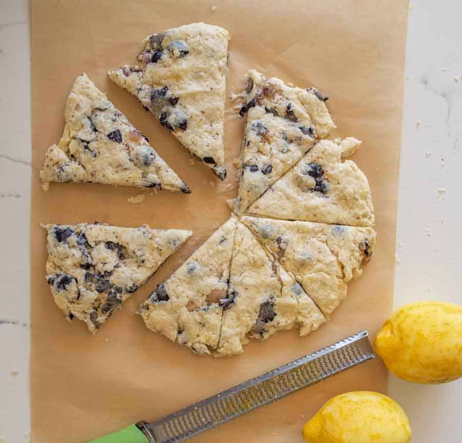 A round, unbaked blueberry scone dough is cut into wedges on parchment paper. A zester and two whole lemons are beside the dough on a white countertop.