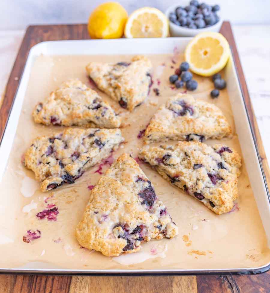A baking sheet with six triangular blueberry scones on parchment paper. Some blueberries and halved lemons are in the background, along with a bowl of fresh blueberries.
