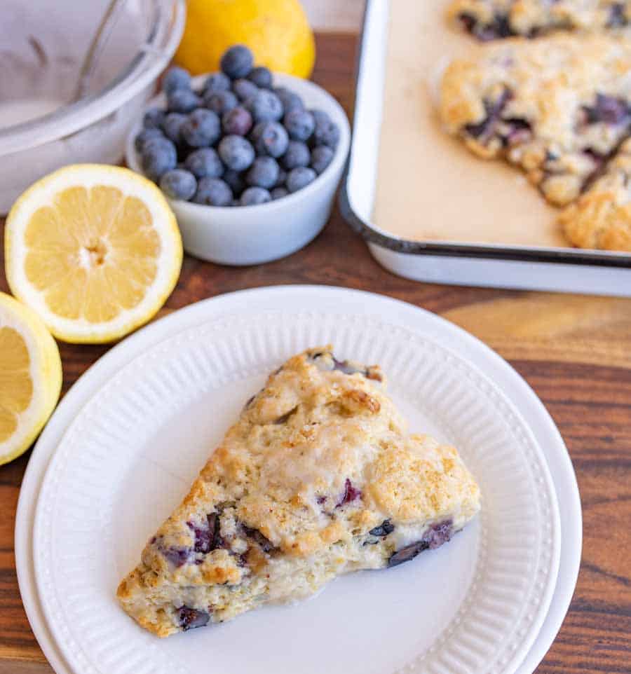 A triangular blueberry scone with glaze sits on a white plate. Nearby are fresh blueberries in a bowl, sliced lemons, and a tray with more scones on a wooden surface.