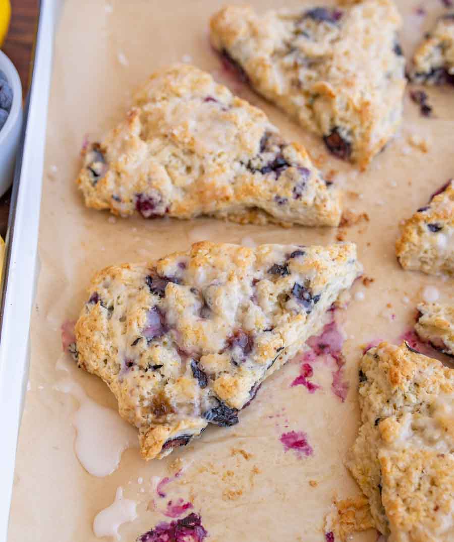 Triangular blueberry scones with a light glaze rest on a baking sheet lined with parchment paper. Some purple berry spots and glaze drips are visible around the baked scones.
