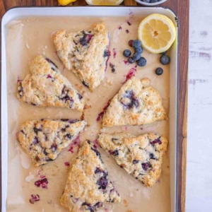 Six triangular blueberry scones are arranged on a parchment-lined baking tray. Beside the tray are fresh blueberries, a whole lemon, and a halved lemon, all placed on a wooden surface.