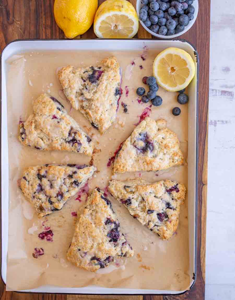 Six triangular blueberry scones are arranged on a parchment-lined baking tray. Beside the tray are fresh blueberries, a whole lemon, and a halved lemon, all placed on a wooden surface.