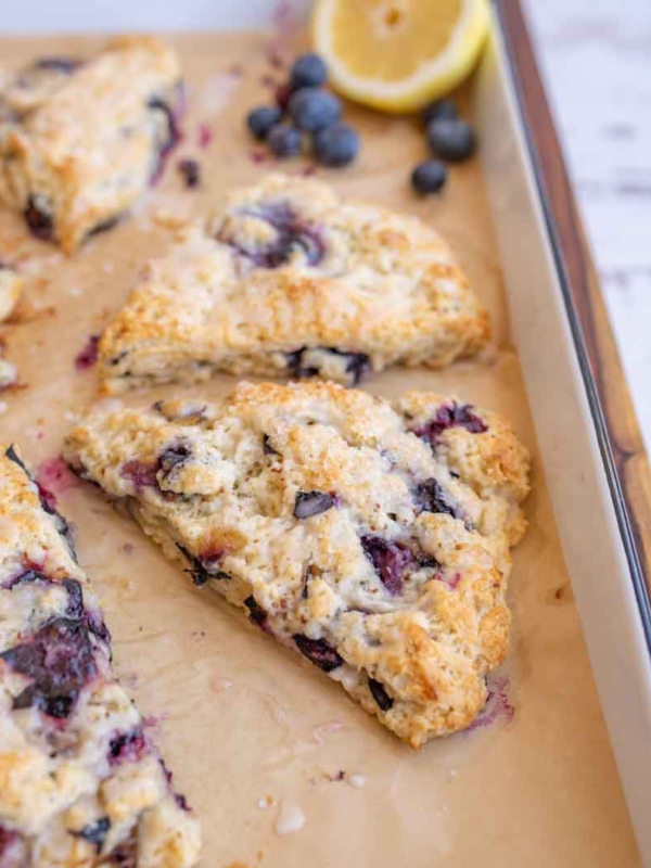 A freshly baked blueberry scone sits on parchment paper, surrounded by more scones, blueberries, and a halved lemon on a baking sheet.