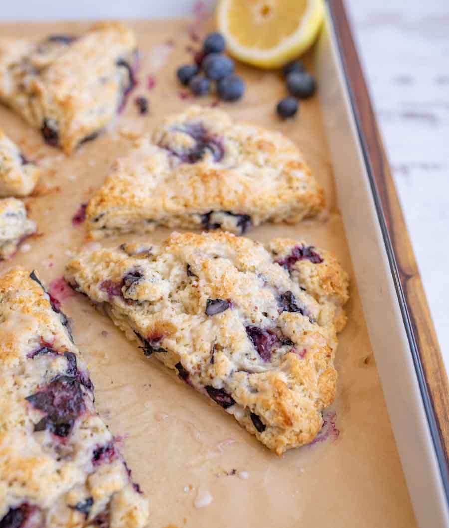 A freshly baked blueberry scone sits on parchment paper, surrounded by more scones, blueberries, and a halved lemon on a baking sheet.