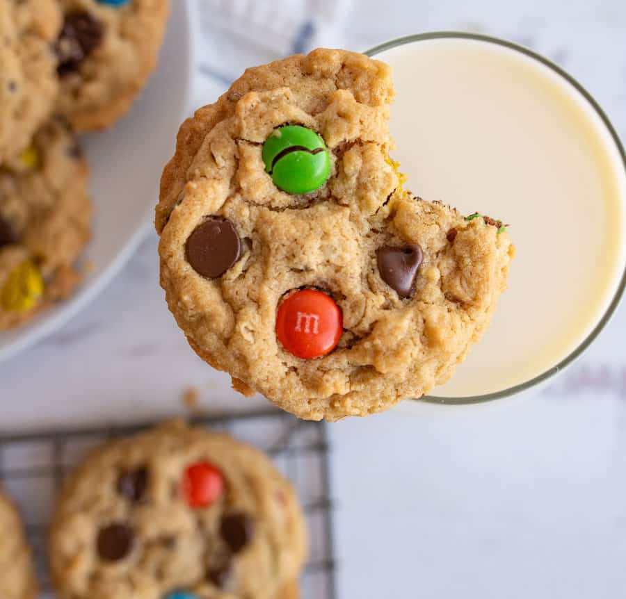 A close-up of a cookie with colorful chocolate candies and a bite taken out, resting on the rim of a glass of milk. More cookies are visible on a plate and a wire rack in the background.