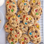 A cooling rack filled with freshly baked cookies containing chocolate chips and colorful candy-coated chocolates, with a checkered cloth underneath.