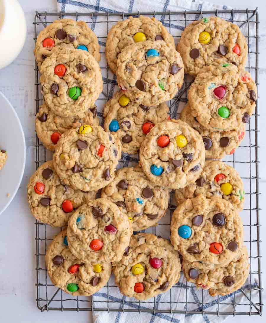 A cooling rack filled with freshly baked cookies containing chocolate chips and colorful candy-coated chocolates, with a checkered cloth underneath.