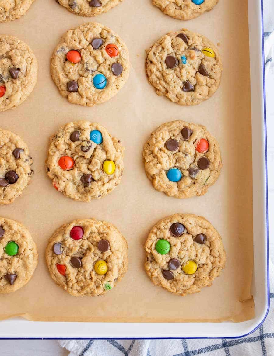 A baking tray lined with parchment paper holds twelve freshly baked cookies with chocolate chips and colorful candy-coated chocolates. The cookies are arranged neatly in rows.