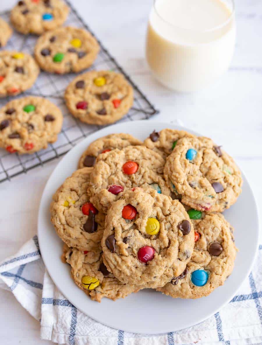 A plate of cookies with colorful chocolate candies and chocolate chips sits on a checkered towel, with more cookies on a cooling rack and a glass of milk in the background.