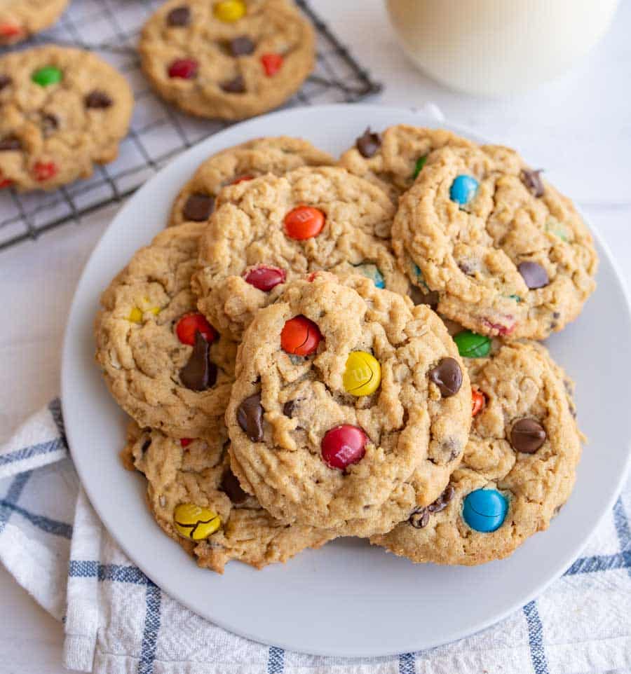 A plate of homemade cookies with colorful candy-coated chocolates and chocolate chips sits on a striped cloth, with more cookies and a glass of milk in the background.
