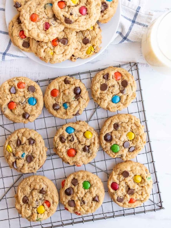 A cooling rack holds oatmeal cookies with colorful candy-coated chocolates and chocolate chips. More cookies are stacked on a plate nearby, with a glass of milk in the corner.